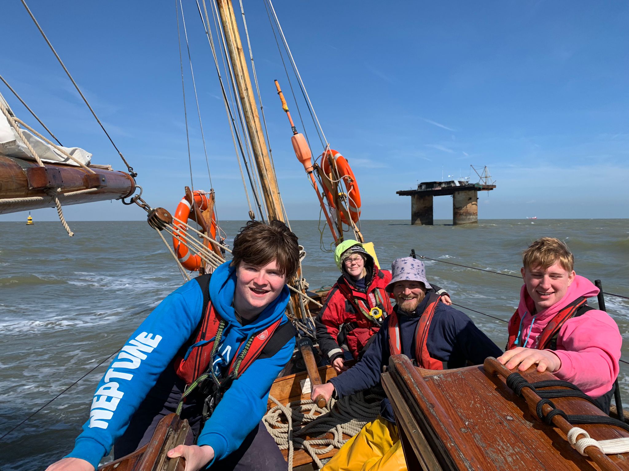 Four people at the stern of Duet, smiling at the camera, one with their hand on the ship's tiller