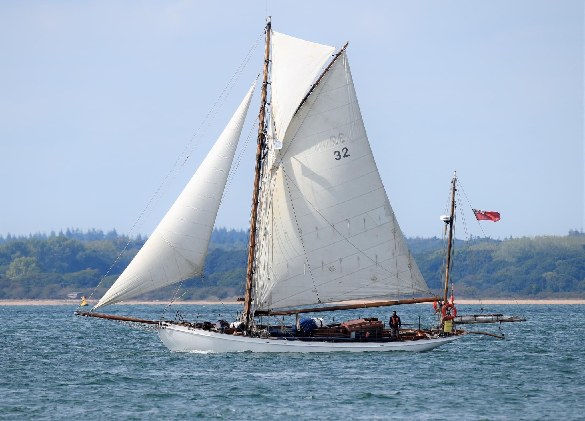 Portside beam view of Duet under sail with forested shore in the background