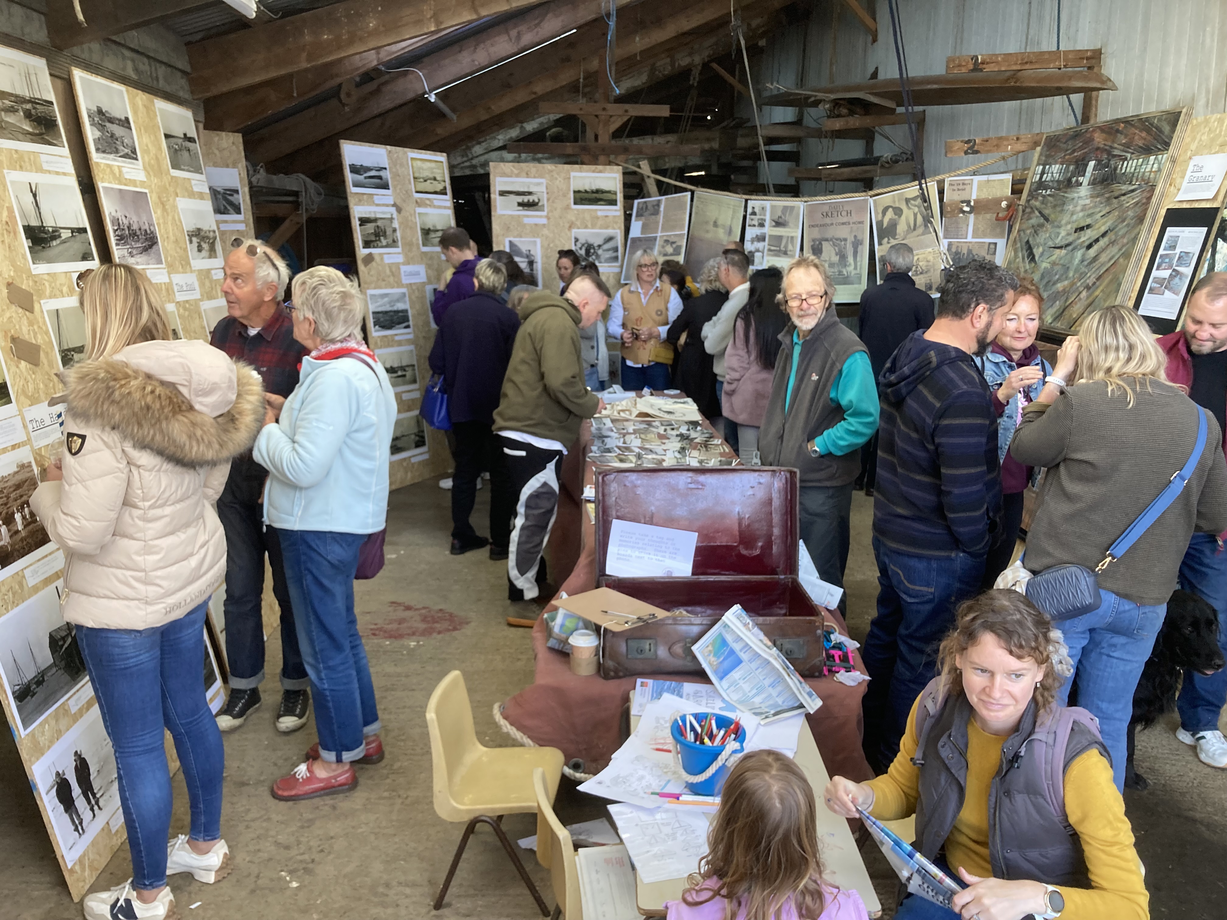 Photo of exhibition space with archival photographs mounted on standing boards. Multiple people are milling about. The Centre of the image has a table with old photographs ready for viewing.