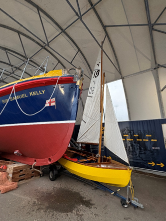 Gull Mark 1 dinghy standing at the port bow of Sir Samuel Kelly. The hull of the dinghy is warm yellow, and the two sails are set