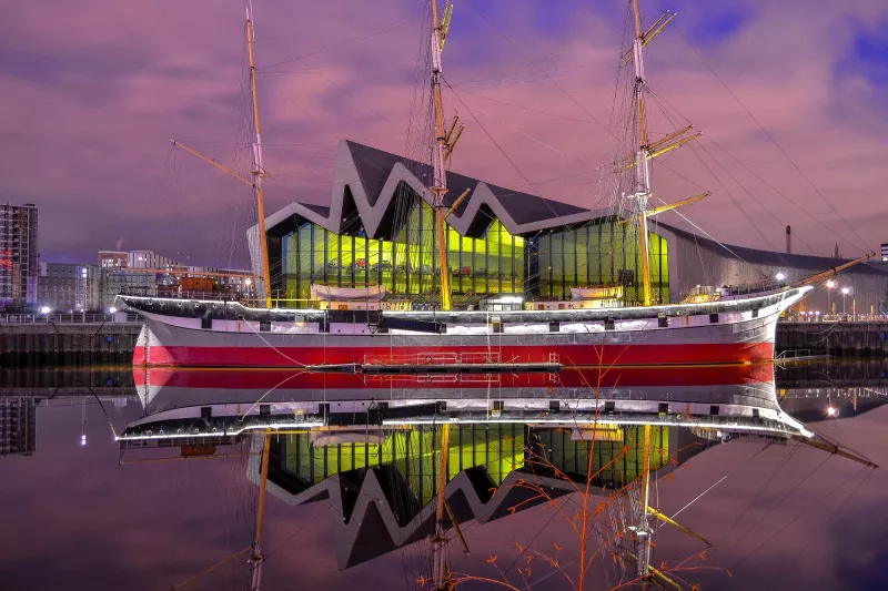 A starboard view of Tall Ship Glenlee. The water and sky are tinted a pinkish purple colour, and the windows in the grey building behind the ship are lit up neon green. The water shows a clear reflection of the ship and the building behind it.