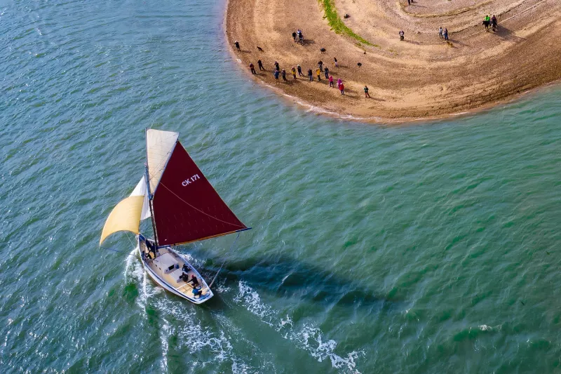 A bird's eye view of CK171 under ful sail against the backdrop of teal coloured water. The ship is in the lower left of the image. The upper right shows a circular edge of land, with multiple people gathered on the beach to observe the ship.