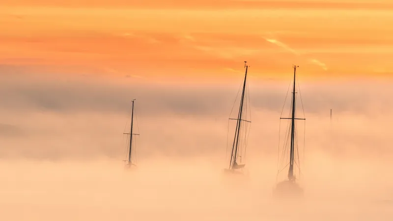 Orange hues cover the whole image. Bottom half of the vista is covered in fog, with three dark masts with stowed sails emerging from the fog in the centre of the photo.