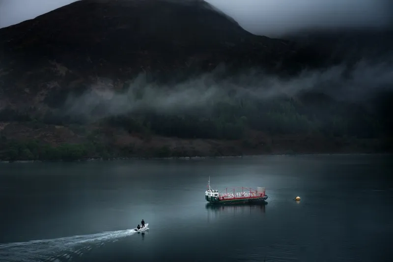 The MV Glenachullish sits in the low centre of the image. A smal white boat with an outboard engine and two people dressed in black approaches the MV from the bottom left corner. The water is dark and there are black towering peaks in the background with a stripe of fog traversing across them.
