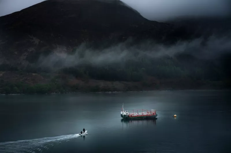 The MV Glenachullish sits in the low centre of the image. A smal white boat with an outboard engine and two people dressed in black approaches the MV from the bottom left corner. The water is dark and there are black towering peaks in the background with a stripe of fog traversing across them.
