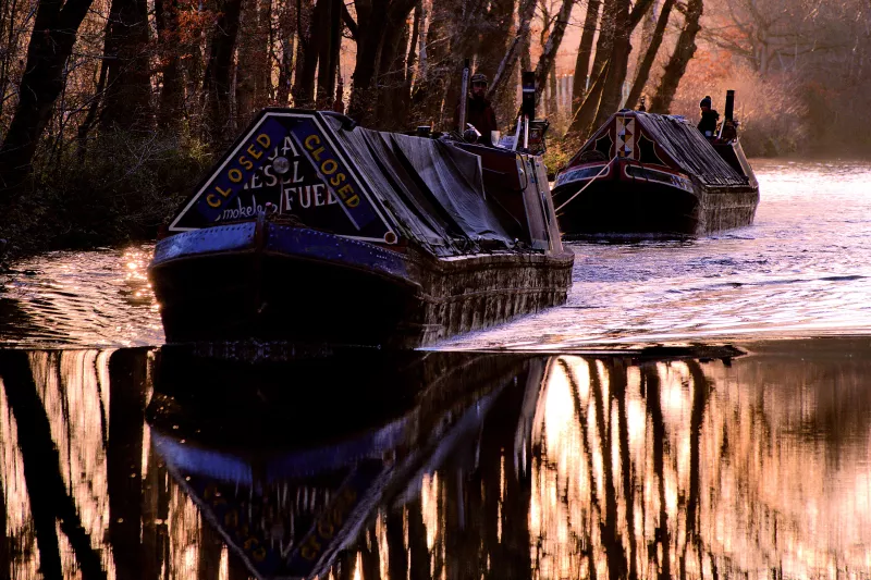 Two canal boats are sailing one behind the other on a crisp autumnal looking morning, with dark trees towering behind them and reflected in the water. 