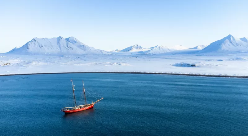 The image is bisected, with open water on the lower half and snowy mountains, ice, and sky in the upper half. A two-masted schooner with a red hull sits in the left foreground part of the image, none of her sails are set.