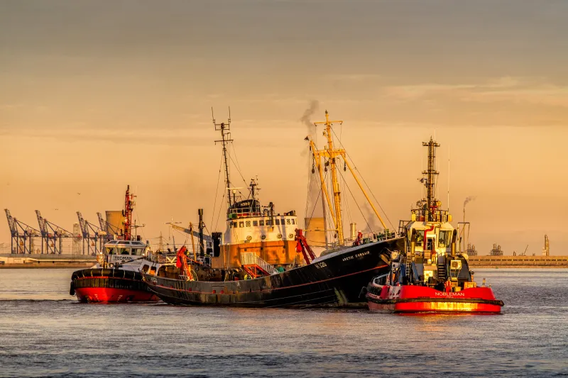 Trawle Arctic Corsair is shown in the centre of the image from her starboard side. She's accompanied by two tugs, Nobleman at the bow and Yeoman at the stern. The yellow hues of the golden hour cast the three ships aglow. Port cranes are standing in the background on the left.