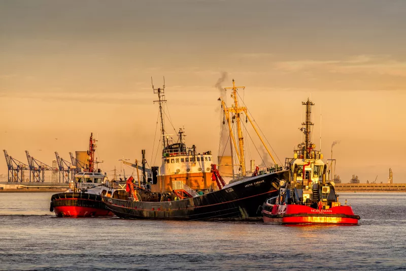Trawle Arctic Corsair is shown in the centre of the image from her starboard side. She's accompanied by two tugs, Nobleman at the bow and Yeoman at the stern. The yellow hues of the golden hour cast the three ships aglow. Port cranes are standing in the background on the left.