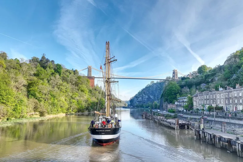 A stern view of Pelican of London with no sails set, going down a river. To the left there is a tall line of trees, to the right a line of old grey slate houses and wooden piers.