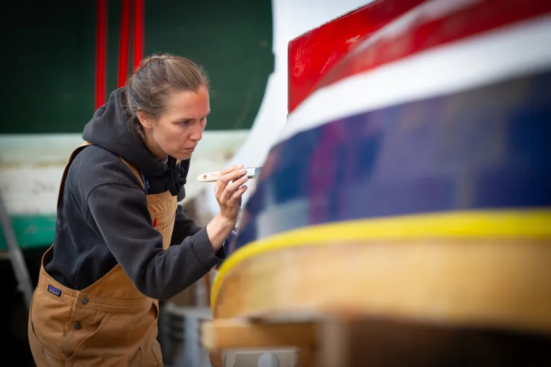 On the right part of an upturned hull of a boat is visible. It has colourful horizontal stripes. A woman on the left of the image is holding a brush to the hull, painting the boat.
