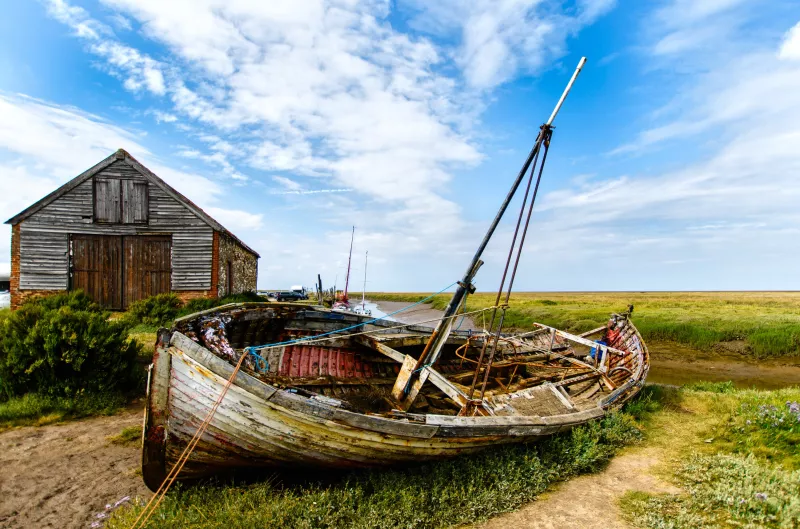 A decrepit small boat with a single mast is lying on its side in the centre of the image on a patch of green grass by a footpath. Behind it on the left stands a wooden barn. The sky above is blue and speckled with white fluffy clouds.