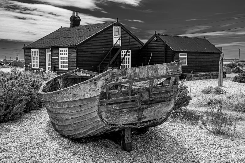 A black and white photo of a decrepit wooden hull sitting on a shingle beach in front of a small wooden cottage.