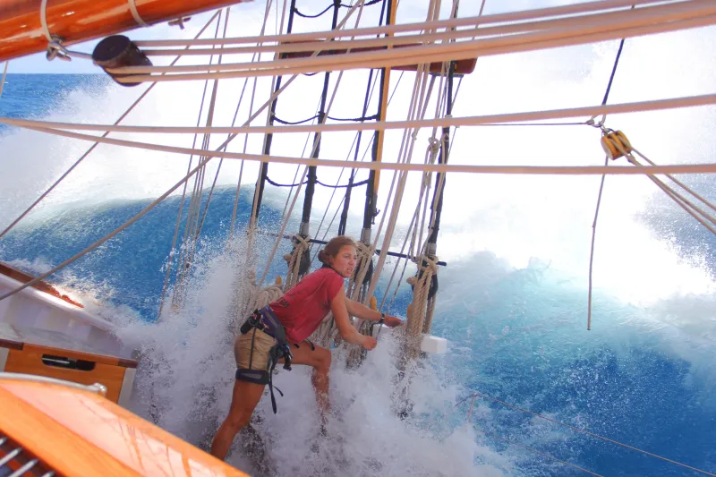 A woman wearing tan shorts and a red t-shirt is standing in the centre of the image in front of a set of ratlines and holding onto a rope. A bright blue wave is pouring onto the deck from behind her and covering her feet in white foam. Ropes and a wooden hatch are in the foreground of the image.