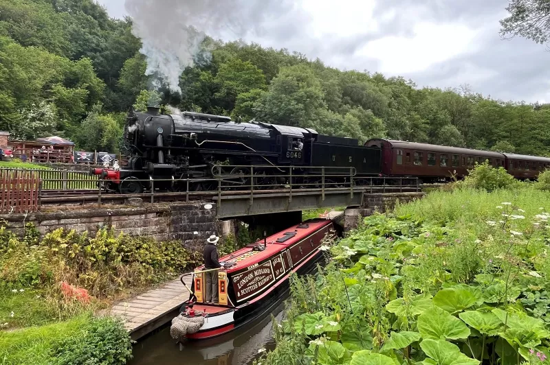 A canal boat and lush vegetation are in the foreground. Above them, a steam train passes on a railway bridge.
