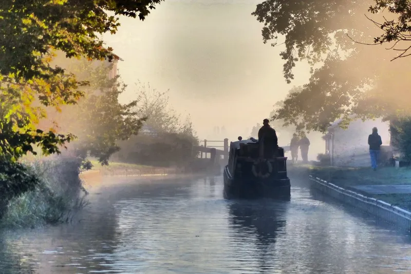 Narrow boat sailing down a canal, trees overhanging the sides and a fog visible in the air