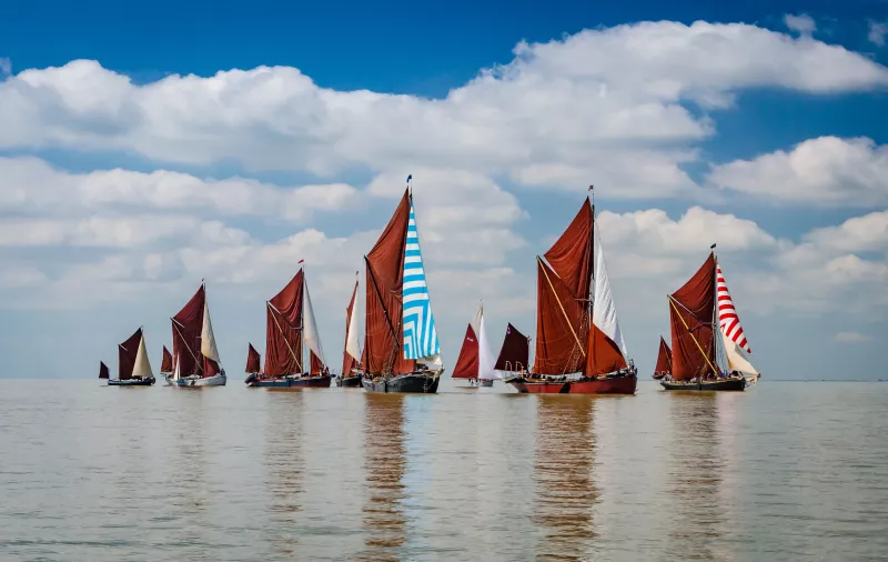 Seven barges in a line under full sail, with a single smack sailing in the background