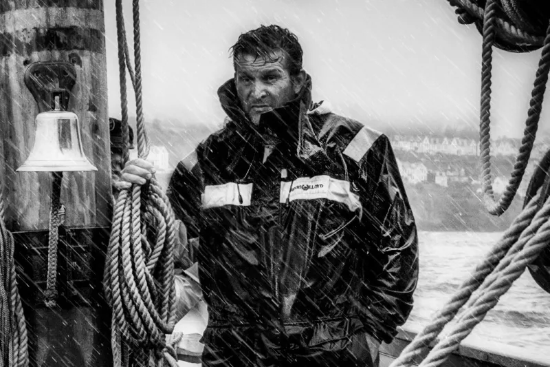 Black and white photo of a sailor standing in the rain in wet weather gear, not looking too happy about it